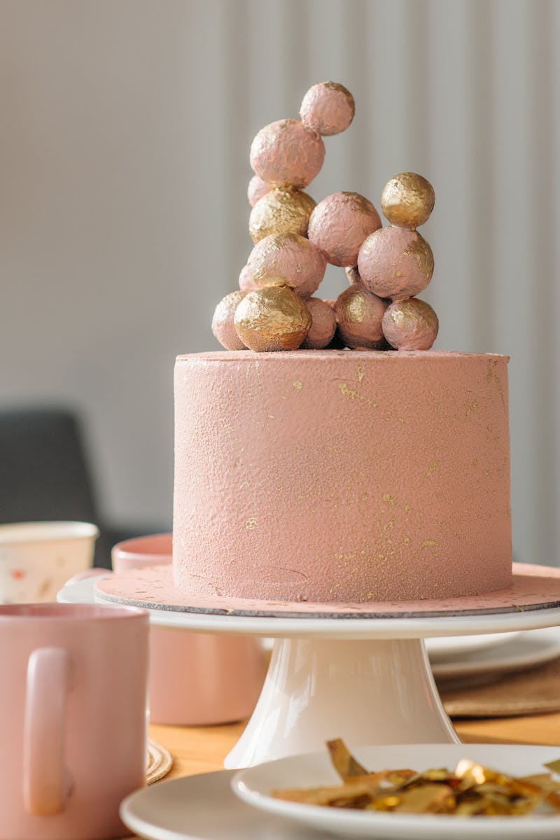 Pink cake topped with gold spheres, elegant design on a table with plates and mugs.